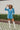Frontal view of the More Than Enough Romper that features a blue corduroy material, a collar neck, a surplice front , a long sleeve with a button, a front zipper, pockets, and pleating on the legs.