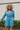 Frontal view of the More Than Enough Romper that features a blue corduroy material, a collar neck, a surplice front , a long sleeve with a button, a front zipper, pockets, and pleating on the legs.