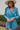 Frontal view of the More Than Enough Romper that features a blue corduroy material, a collar neck, a surplice front , a long sleeve with a button, a front zipper, pockets, and pleating on the legs.