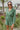 Frontal view of the More Than Enough Romper that features a green corduroy material, a collar neck, a surplice front, a long sleeve with a button, a front zipper, pockets, and pleating on the legs.