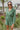 Frontal view of the More Than Enough Romper that features a green corduroy material, a collar neck, a surplice front, a long sleeve with a button, a front zipper, pockets, and pleating on the legs.