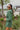 Side view of the More Than Enough Romper that features a green corduroy material, a collar neck, a surplice front, a long sleeve with a button, a front zipper, pockets, and pleating on the legs.