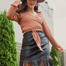Front view of model wearing the Backstage Pass Skirt features a black faux-leather material, a thick elastic waist band, a high-waisted fit, fringe detailing along the bottom hem, and a mini length. Worn with brown top.