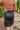 Close-up back view of model wearing the Backstage Pass Skirt features a black faux-leather material, a thick elastic waist band, a high-waisted fit, fringe detailing along the bottom hem, and a mini length. Worn with brown top.