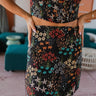 Frontal view of the Everglow Mini Skirt that features a black colored material, a multi-colored floral print, a high-rise waist, a side slit, and a side zipper closure.