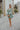 Full body view of model wearing the Forever Young Floral Dress that has a sage green, blue, white and metallic gold floral print, long balloon sleeves with elastic wrists, a lined skirt with a ruffled hemline, and a back zipper.
