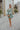 Full body view of model wearing the Forever Young Floral Dress that has a sage green, blue, white and metallic gold floral print, long balloon sleeves with elastic wrists, a lined skirt with a ruffled hemline, and a back zipper.