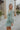 Side view of model wearing the Forever Young Floral Dress that has a sage green, blue, white and metallic gold floral print, long balloon sleeves with elastic wrists, a lined skirt with a ruffled hemline, and a back zipper.
