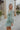 Side view of model wearing the Forever Young Floral Dress that has a sage green, blue, white and metallic gold floral print, long balloon sleeves with elastic wrists, a lined skirt with a ruffled hemline, and a back zipper.