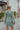 Back view of model wearing the Forever Young Floral Dress that has a sage green, blue, white and metallic gold floral print, long balloon sleeves with elastic wrists, a lined skirt with a ruffled hemline, and a back zipper.