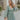 Front view of model wearing the Forever Young Floral Dress that has a sage green, blue, white and metallic gold floral print, long balloon sleeves with elastic wrists, a lined skirt with a ruffled hemline, and a back zipper.