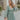 Front view of model wearing the Forever Young Floral Dress that has a sage green, blue, white and metallic gold floral print, long balloon sleeves with elastic wrists, a lined skirt with a ruffled hemline, and a back zipper.