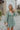 Front view of model wearing the Forever Young Floral Dress that has a sage green, blue, white and metallic gold floral print, long balloon sleeves with elastic wrists, a lined skirt with a ruffled hemline, and a back zipper.