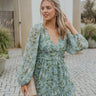 Front view of model wearing the Forever Young Floral Dress that has a sage green, blue, white and metallic gold floral print, long balloon sleeves with elastic wrists, a lined skirt with a ruffled hemline, and a back zipper.