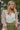 Front view of model wearing the Someday Soon Bodysuit that has cream satin fabric with a surplice neckline, a knit thong bottom with snap closures, and organza puff sleeves. Worn under green skirt.