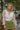 Front view of model wearing the Someday Soon Bodysuit that has cream satin fabric with a surplice neckline, a knit thong bottom with snap closures, and organza puff sleeves. Worn under green skirt.