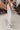 Side view of model wearing the Most Wanted Jeans, that have white denim, a crossover waist, a high-rise fit, a button closure, two front pockets, distressing at the knees, two back pockets, and a straight leg fit.