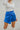 Close front view of model wearing the Bright Idea Skort, that has royal blue satin fabric, a front skirt overlay with a twist detail, and a back zipper