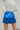 Back view of model wearing the Bright Idea Skort, that has royal blue satin fabric, a front skirt overlay with a twist detail, and a back zipper