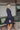 Side view of the On The Spot Mini Dress that features a navy colored material, a deep V neckline, a long cuffed sleeve, side cut-outs, a tiered design, a back tie closure, an open back, and a mini length.