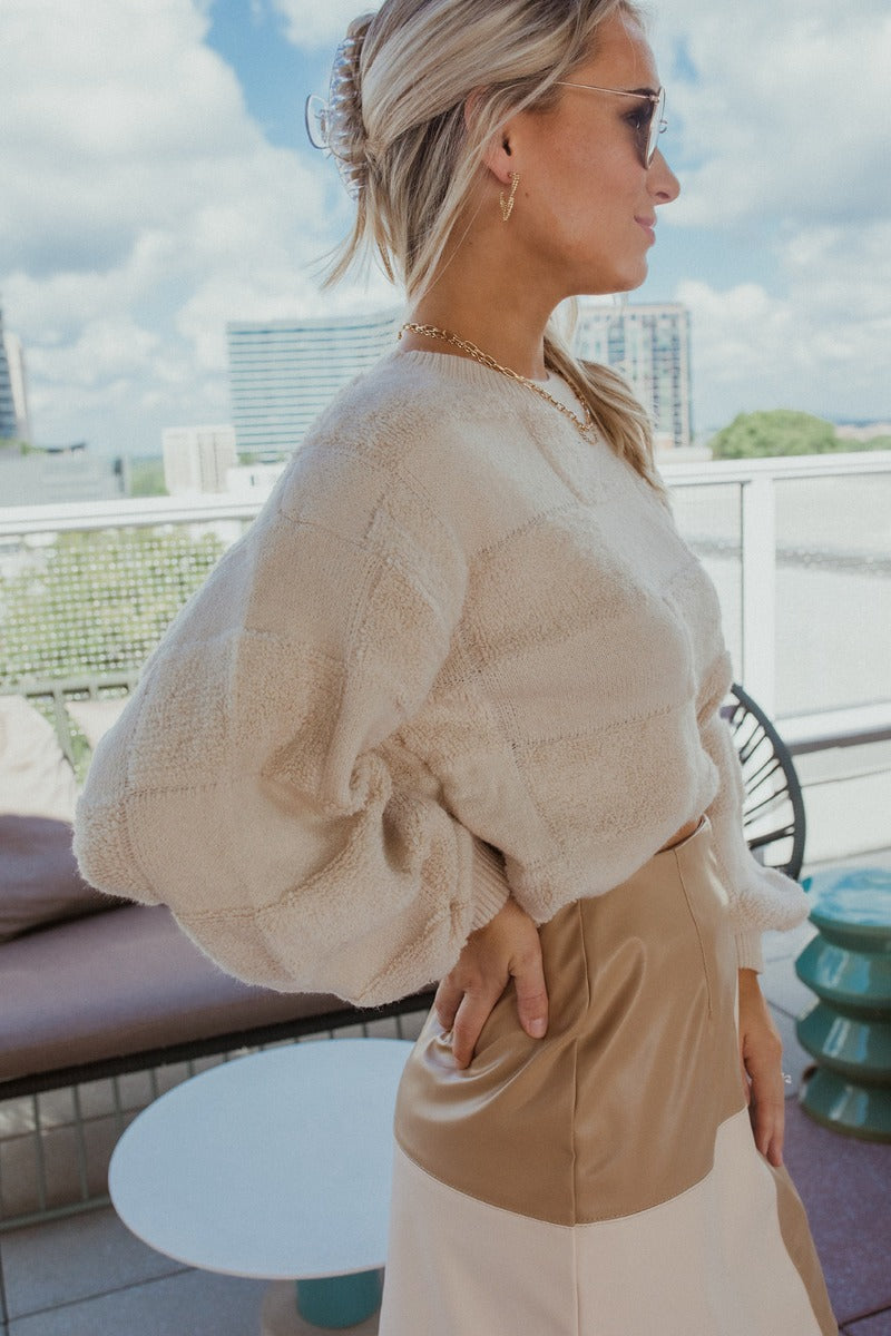 Side view of the Blank Check Sweater that features a cream colored knit material, a round neck, a long sleeve, and a patchwork textured design.