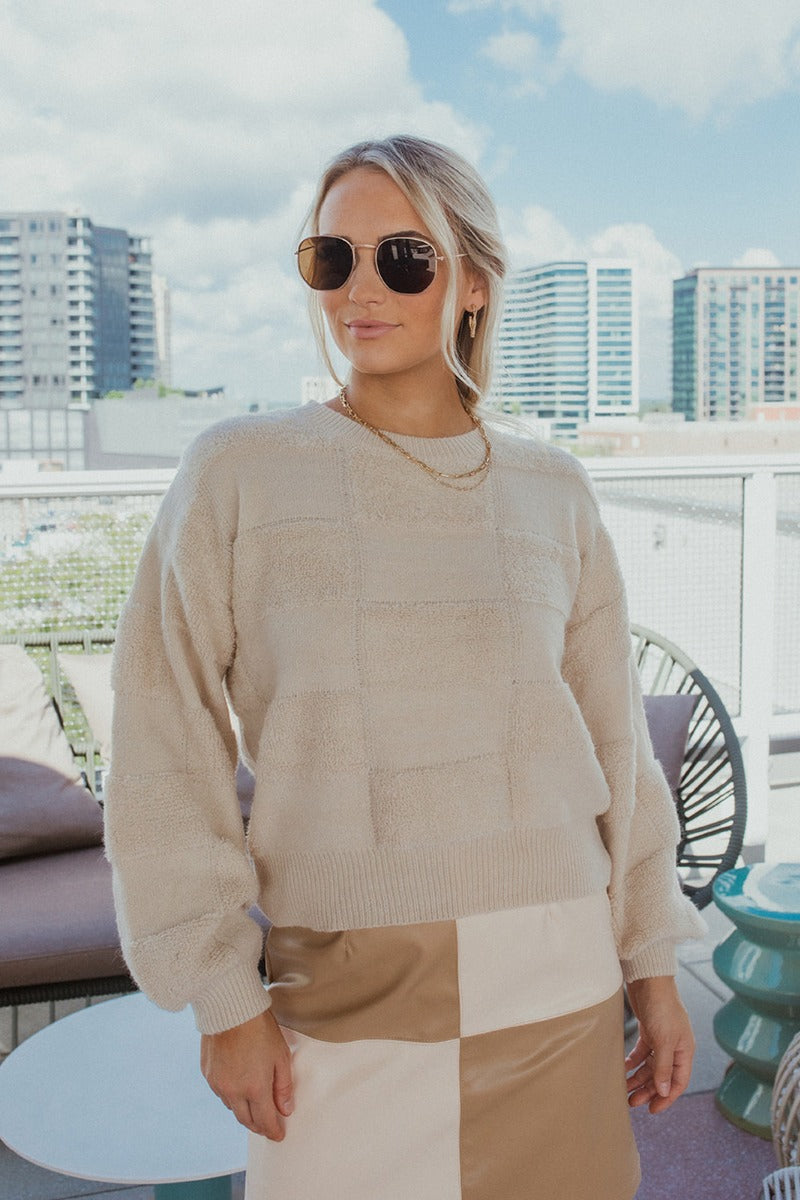 Frontal view of the Blank Check Sweater that features a cream colored knit material, a round neck, a long sleeve, and a patchwork textured design.