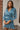 Frontal view of the Every Breath Mini Dress features a blue shimmer material, a surplice neckline, long sleeves with smocked detailing at the wrist, a front ruched detail, a ruched back seam, a back zipper closure, an open back with a button closure, and 