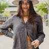 Frontal view of the Bare With Me Top that features a charcoal grey gauze material, a V neckline, a button-up front, a long cuffed sleeve, raw hemlines through out, and side pockets.