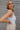 Side view of model wearing the Play It Cool Bodysuit that has white ribbed fabric, a v-neckline, a sleeveless body, a thong bottom and hook closures.