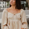 Frontal view of the love You More Top that features a champagne colored silk material, a straight neckline with a tie detail, a long sleeve, a pleated front, a smocked back, a back zipper closure, and a flowy fit.