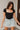 Front view of model wearing the Nothing Better Bodysuit, that has black stretchy ribbed fabric, a square neckline, short sleeves, and a thong bottom with hook and eye closures. Worn with denim shorts.