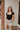 Front view of model wearing the Nothing Better Bodysuit, that has black stretchy ribbed fabric, a square neckline, short sleeves, and a thong bottom with hook and eye closures. Worn with denim shorts.
