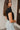 Side view of model wearing the Nothing Better Bodysuit, that has black stretchy ribbed fabric, a square neckline, short sleeves, and a thong bottom with hook and eye closures. Worn with denim shorts.