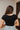 Back view of model wearing the Nothing Better Bodysuit, that has black stretchy ribbed fabric, a square neckline, short sleeves, and a thong bottom with hook and eye closures. Worn with denim shorts.
