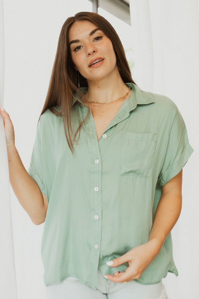 Front view of model wearing the Can't Go Wrong Top, which has a sage green colored material, a collar neck, a button-up front, a short cuffed sleeve, a front pocket, and a flowy fit