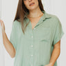 Front view of model wearing the Can't Go Wrong Top, which has a sage green colored material, a collar neck, a button-up front, a short cuffed sleeve, a front pocket, and a flowy fit