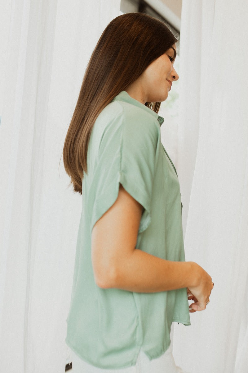Side view of model wearing the Can't Go Wrong Top, which has a sage green colored material, a collar neck, a button-up front, a short cuffed sleeve, a front pocket, and a flowy fit
