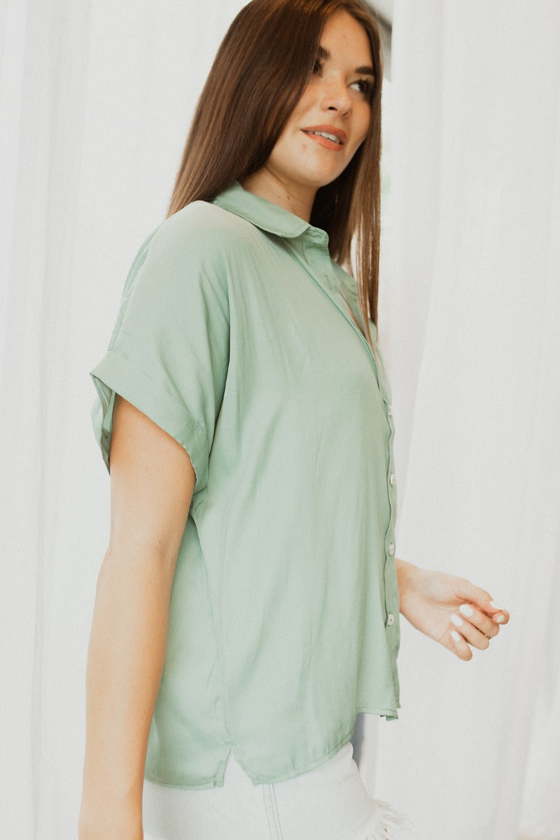 Side view of model wearing the Can't Go Wrong Top, which has a sage green colored material, a collar neck, a button-up front, a short cuffed sleeve, a front pocket, and a flowy fit