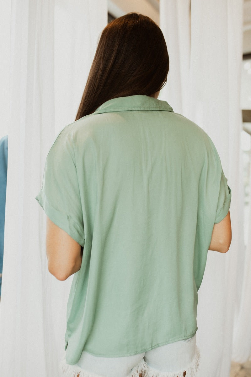 Back view of model wearing the Can't Go Wrong Top, which has a sage green colored material, a collar neck, a button-up front, a short cuffed sleeve, a front pocket, and a flowy fit