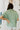 Back view of model wearing the Can't Go Wrong Top, which has a sage green colored material, a collar neck, a button-up front, a short cuffed sleeve, a front pocket, and a flowy fit