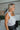 Side view of model wearing The Angel Baby Feather Top features a white silk material, a sleeveless design with thin straps, feather detailing on the straps, a cowl neckline, and a flowy fit.