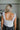 Back view of model wearing The Angel Baby Feather Top features a white silk material, a sleeveless design with thin straps, feather detailing on the straps, a cowl neckline, and a flowy fit.