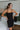 Front view of model wearing The Starlight Rhinestone Fringe Dress features a black colored material, a straight neckline, a sleeveless design with thin adjustable straps, rhinestone fringe detailing at the bottom side hem, criss cross straps in the back, 