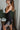 Side view of model wearing The Starlight Rhinestone Fringe Dress features a black colored material, a straight neckline, a sleeveless design with thin adjustable straps, rhinestone fringe detailing at the bottom side hem, criss cross straps in the back, a