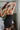 Back view of model wearing The Starlight Rhinestone Fringe Dress features a black colored material, a straight neckline, a sleeveless design with thin adjustable straps, rhinestone fringe detailing at the bottom side hem, criss cross straps in the back, a