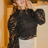 Front view of model wearing The Parisian Wonder Top features a black crochet material, a high round neckline, long sheer sleeves, lace detail at the neckline, a lining underneath, and a back zipper closure.