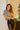 Frontal view of the Timeless Top that features a black colored material, a white houndstooth pattern, short sleeves, gold button detailing at the shoulder, and fringe detailing at the hems.