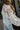 Side view of model wearing The Queen Anne's Lace Top In White features a white colored sheer material, a collar neck, a button-up front, long puffy sleeves, and a tank top liner.