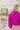 Back view of model wearing the Take Me Out Top, which features a purple colored material, a collar V neck with a button, a long flowy sleeve with a smocking at the wrist, a draped bottom hem. Tucked into beige skirt.