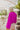 Back view of model wearing the Take Me Out Top, which features a purple colored material, a collar V neck with a button, a long flowy sleeve with a smocking at the wrist, a draped bottom hem. Tucked into beige skirt.